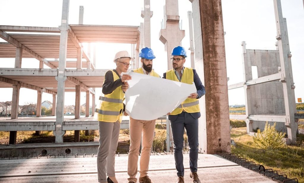 Na zdjęciu widzimy trzech ludzi ubranych w kamizelki odblaskowe i kaski ochronne, stojących na placu budowy. Wspólnie analizują duży plan architektoniczny, być może omawiają szczegóły konstrukcji. W tle widoczna jest częściowo wzniesiona konstrukcja, składająca się z betonowych belek i kolumn.